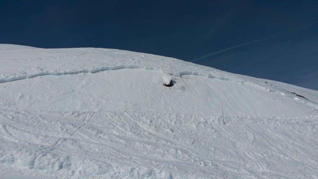Vue d'une pente enneigée avec des stries et une fissure visible dans la neige sous un ciel bleu.