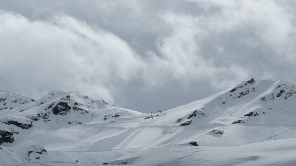 Vue panoramique des montagnes enneigées sous un ciel nuageux, avec des traces de ski visibles sur la neige.