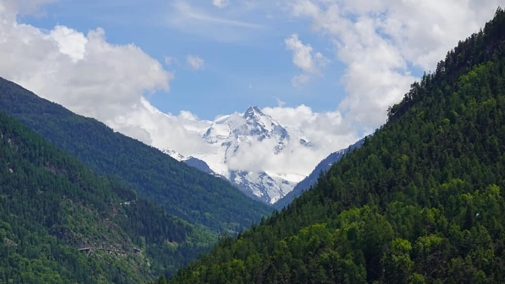 Vue panoramique des montagnes alpines avec des sommets enneigés, entourés de forêts verdoyantes et d'un ciel partiellement nuageux.