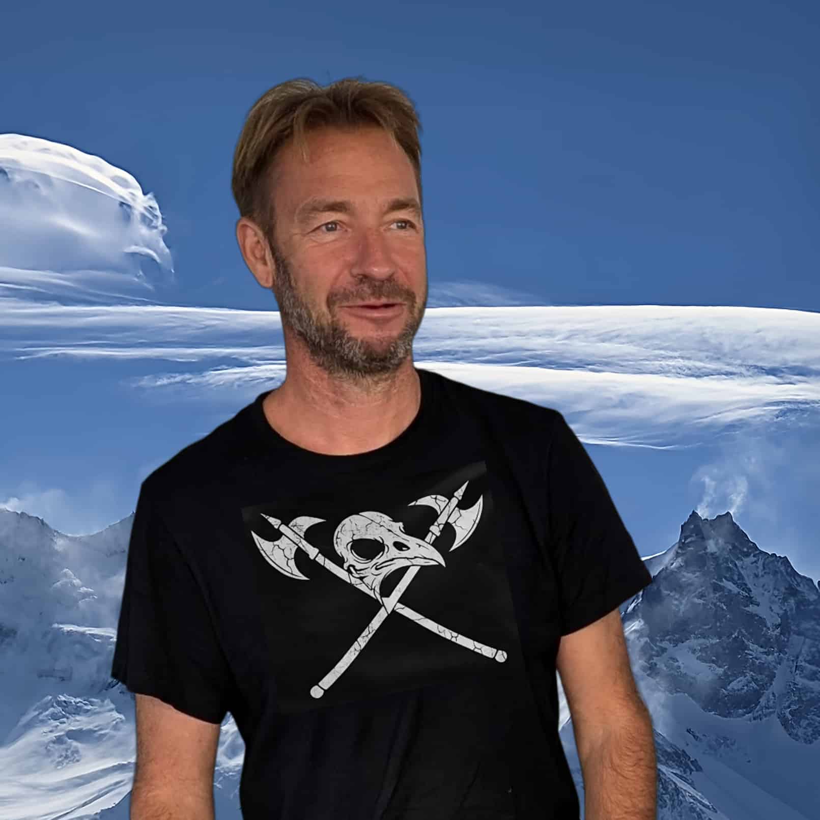 Portrait d'un homme souriant, vêtu d'un t-shirt noir avec un motif graphique, devant un paysage de montagnes et de nuages.