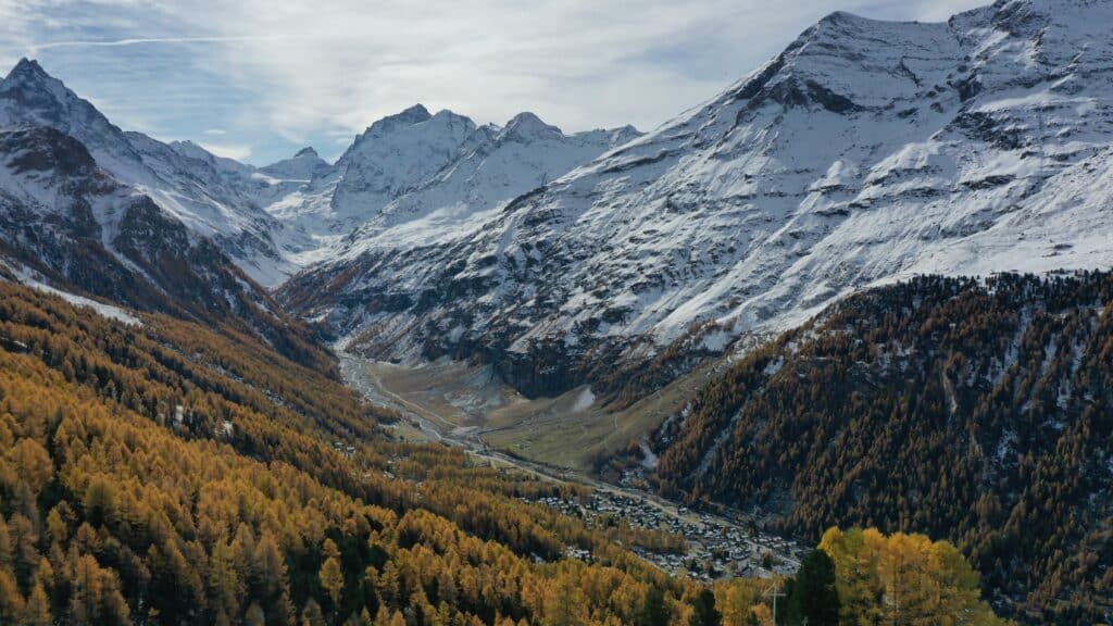 Vue panoramique des montagnes enneigées et des arbres aux feuilles dorées, avec un village visible dans la vallée en bas.