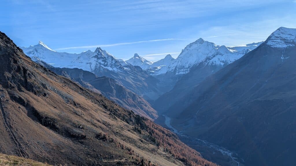 Vue panoramique des montagnes avec des sommets enneigés, un vallon verdoyant et des arbres d'automne dans la vallée.