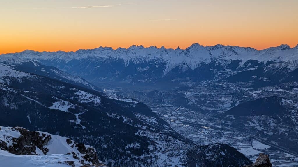 Vue panoramique de montagnes enneigées au coucher du soleil, avec un ciel orange et des vallées environnantes.