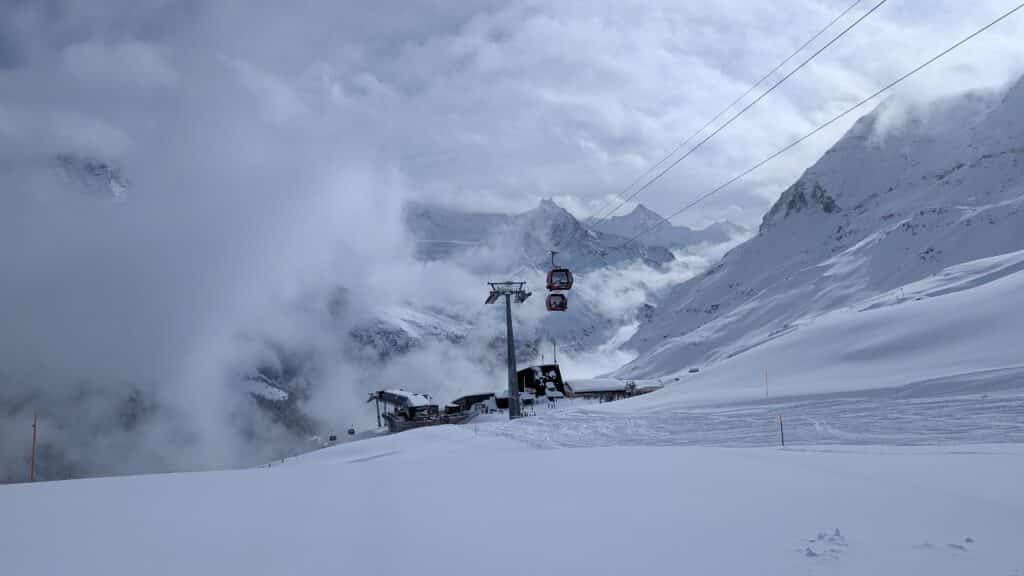 Vue d'une station de ski enneigée avec une télécabine dans un paysage montagneux partiellement nuageux.