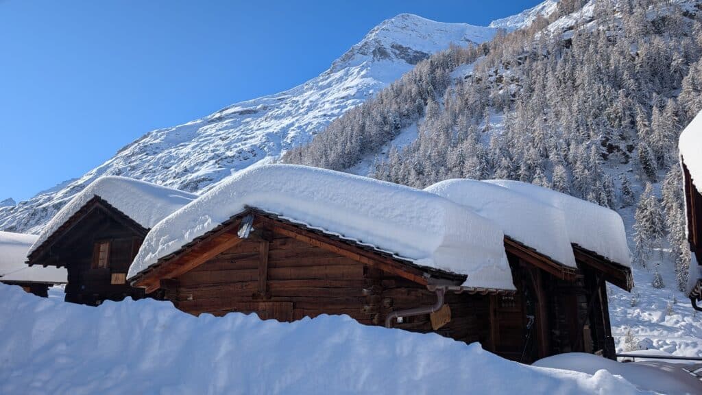 Des chalets en bois recouverts de neige sous un ciel bleu, avec des montagnes enneigées en arrière-plan.
