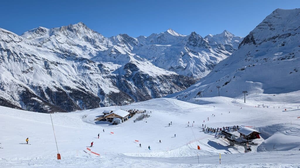 Vue panoramique d'une station de ski avec des skieurs sur des pistes enneigées, entourée de montagnes majestueuses.