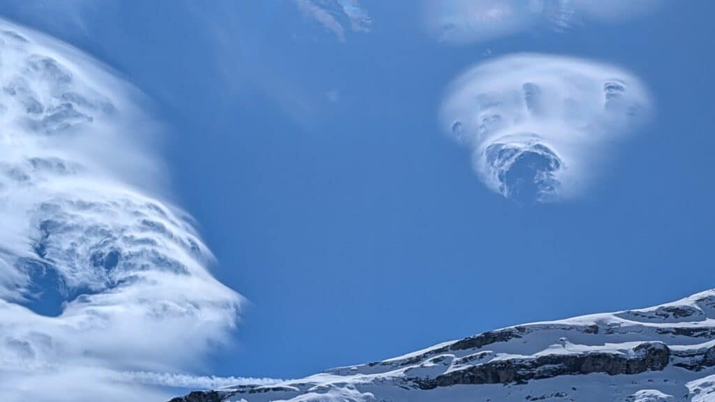 Ciel bleu avec des nuages en forme de lentille et des sommets enneigés en bas de l'image.