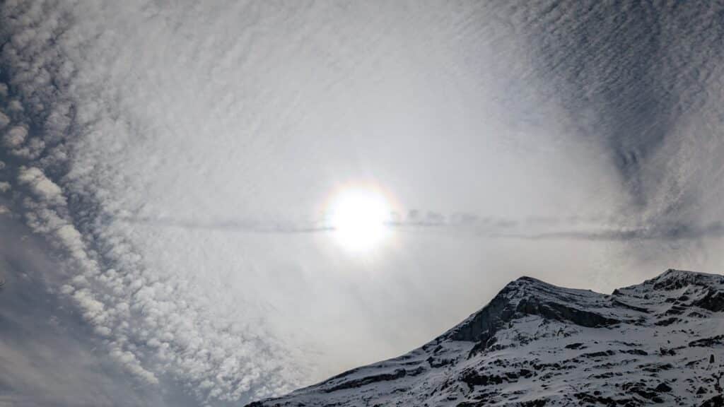 Vue d'un sommet enneigé avec le soleil brillant au-dessus et un ciel partiellement nuageux.