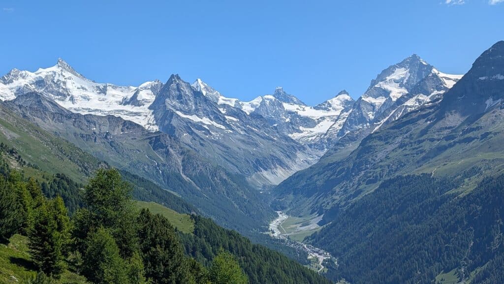 Vue panoramique des montagnes avec sommets enneigés sous un ciel bleu, entourées de verdure au printemps.
