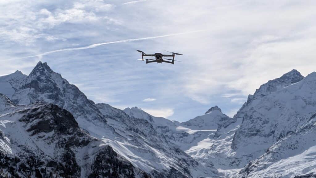 Un drone survole des montagnes enneigées sous un ciel partiellement nuageux.