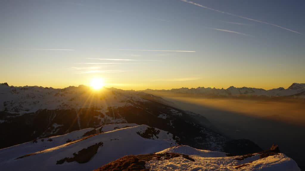 Lever de soleil sur les montagnes enneigées avec des nuages dans la vallée, créant une atmosphère paisible.