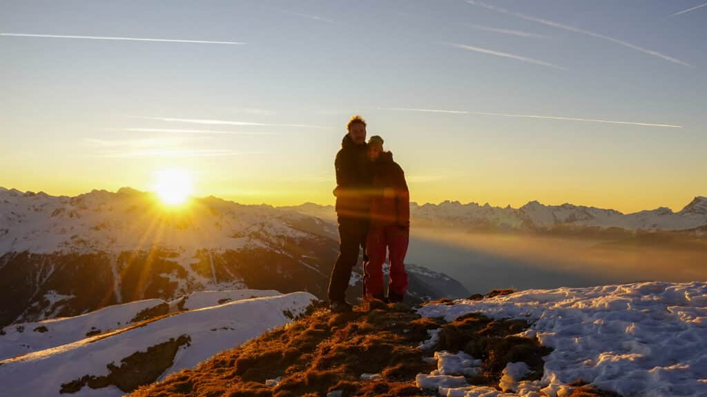 Un couple se tenant par la taille au sommet d'une montagne, avec le soleil se levant derrière eux, illuminant le paysage alpin enneigé.