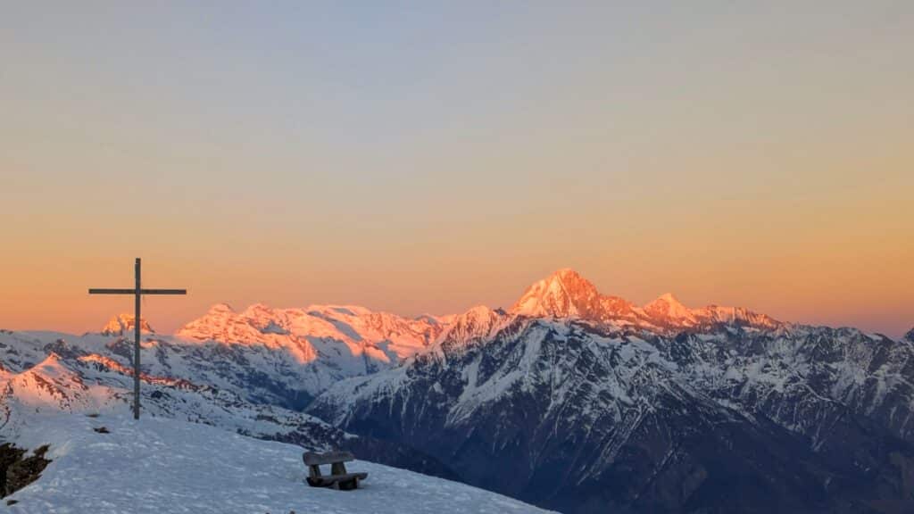 Vue panoramique des montagnes enneigées au coucher du soleil, avec une croix en bois et un banc en premier plan.