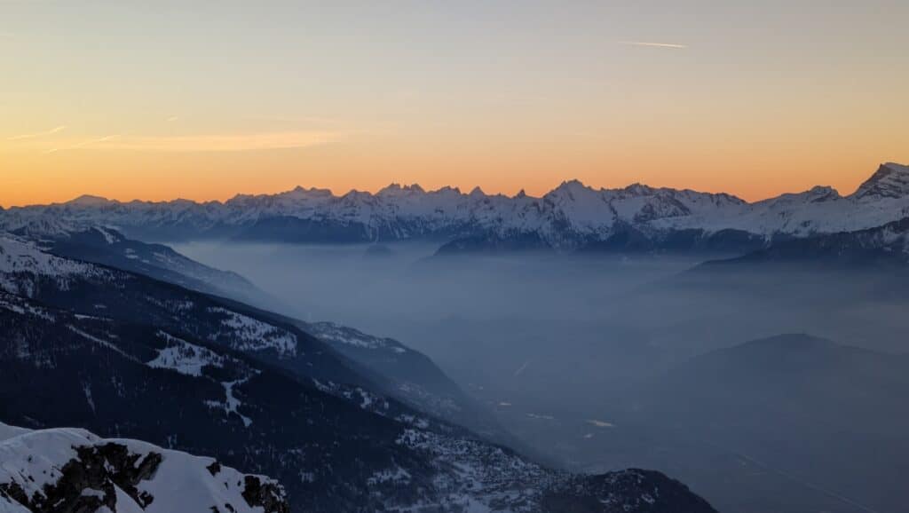 Vue panoramique des montagnes enneigées au crépuscule, avec un ciel aux teintes orange et bleu, et une vallée brumeuse en dessous.