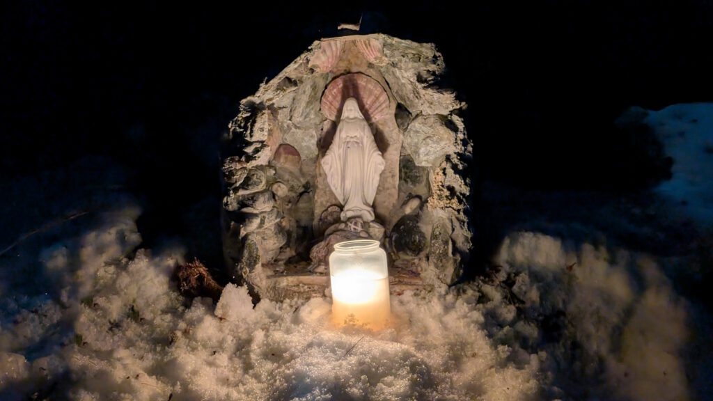 Une statue de la Vierge Marie éclairée par une bougie, entourée de neige et dans un environnement sombre.