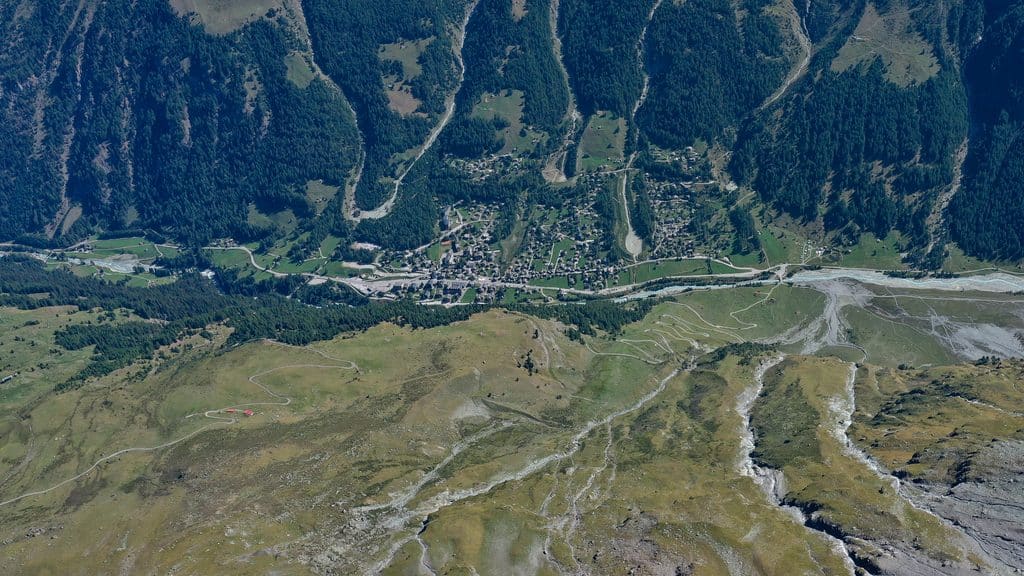 Vue aérienne d'une vallée alpine avec village, forêt, rivière et paraglider.