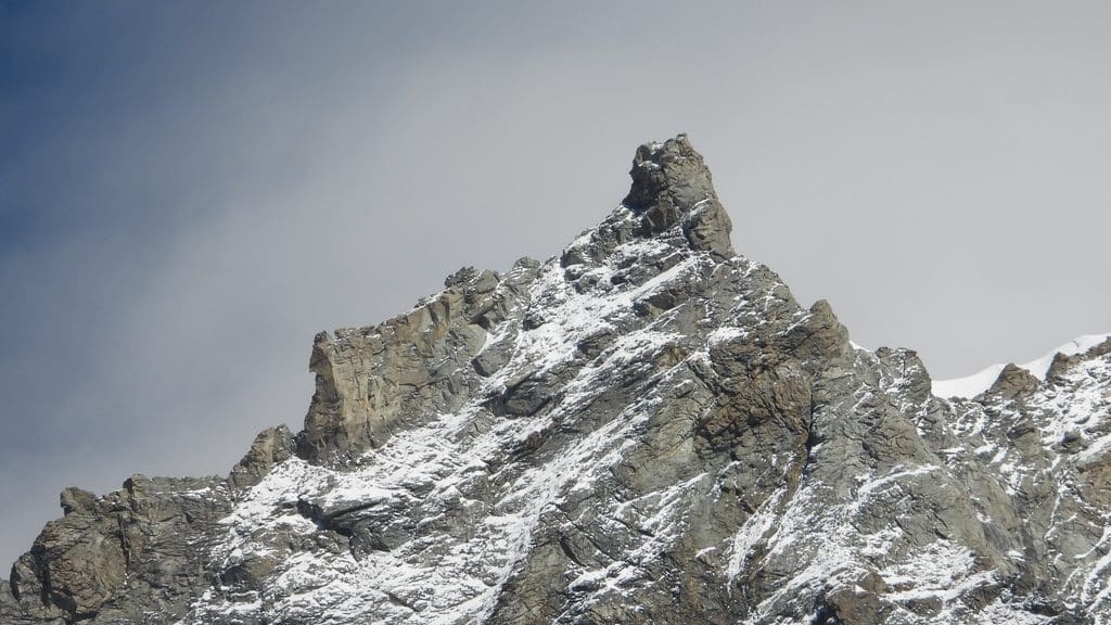 Pic montagneux rocheux et enneigé sous un ciel nuageux.