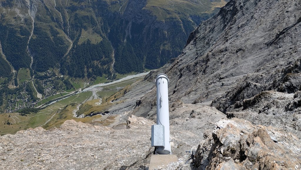 Instrument blanc sur pente rocheuse, vallée alpine en contrebas.